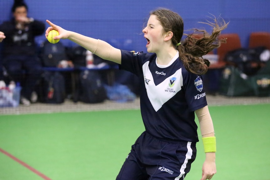 Holding the ball in her right hand and wearing a Victoria kit, Elise Collier yells and points towards the umpire.