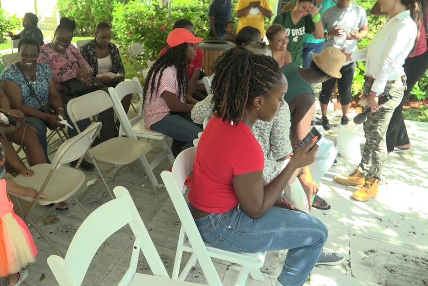 A group of people, some of them looking at their phones, sit on rows of chairs.