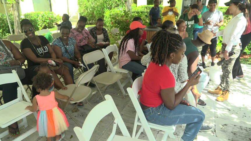 A group of people, some of them looking at their phones, sit on rows of chairs.