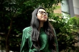 A woman wearing glasses stands in front of a tree.