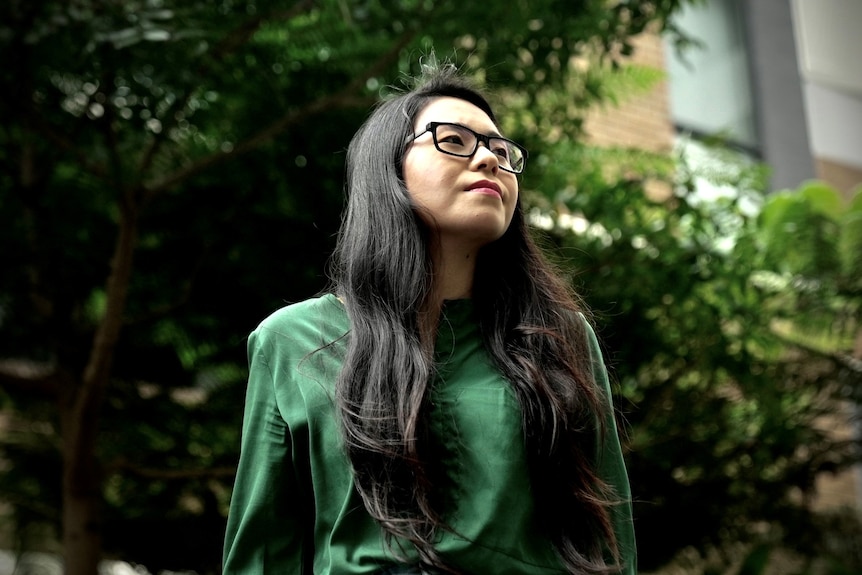 Una mujer con gafas se para frente a un árbol.