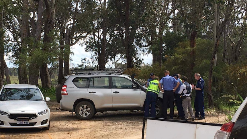 Police officers cluster around a car bonnet, in a bushland area.
