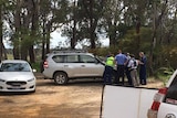 Police officers cluster around a car bonnet, in a bushland area.