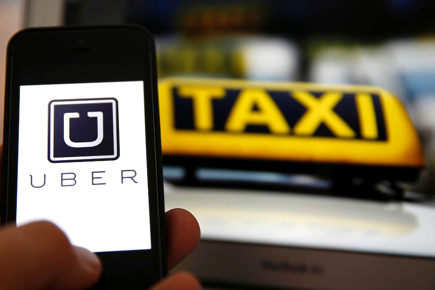 The Uber logo is seen on a smartphone screen in front of a taxi sign.