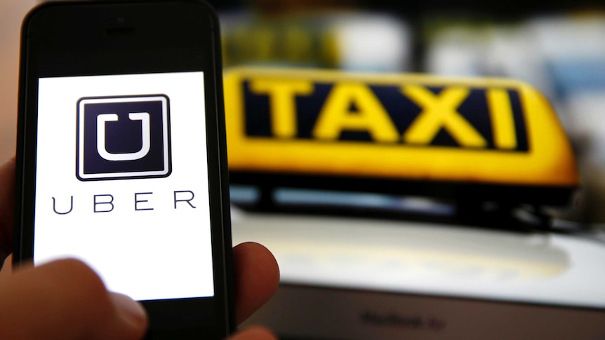 The Uber logo is seen on a smartphone screen in front of a taxi sign.