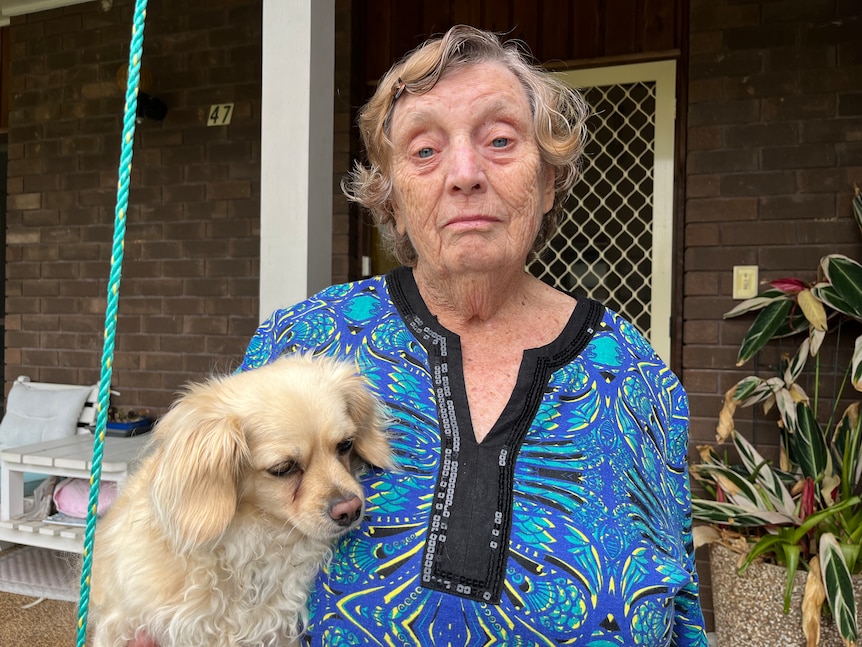 An older woman holding a dog stands in front of her house, looking glum.