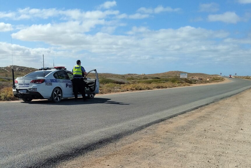 A wide shot of a police car and police officer parked on a bitumen road.