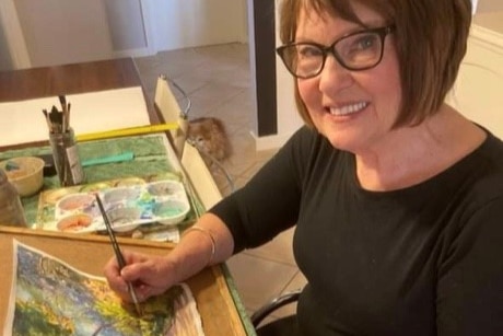 Woman wearing glasses looking at camera, with brush in hand and painting on desk
