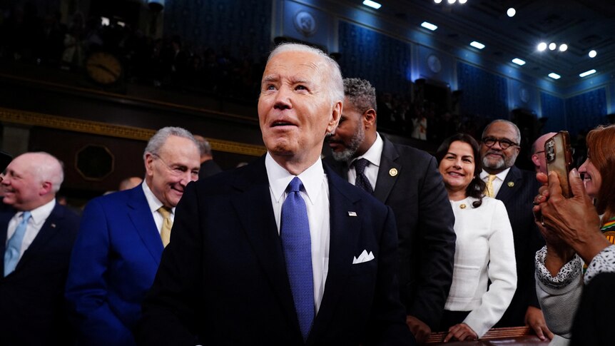 Joe Biden wearing a blue tie with his suit stands in a crowd of people.