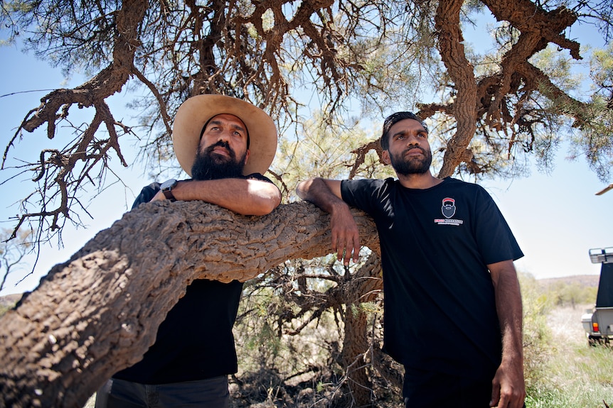 Two men in black t-shirts sand against a tree trunk. 