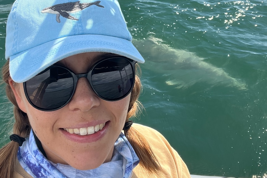 A smiling woman wearing sunglasses with a dolphin in the water behind her.