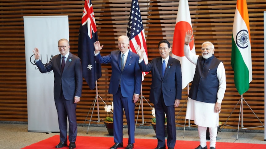Four men in suits smile and wave at cameras with four flags behind them.