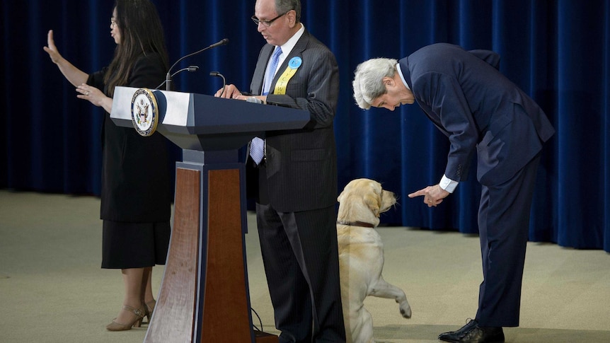 John Kerry bends over and points at his dog behind a man speaking at a podium.