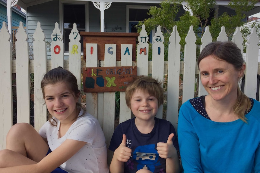 Two children and their mother sit against a white picket fence that says 'Origami'