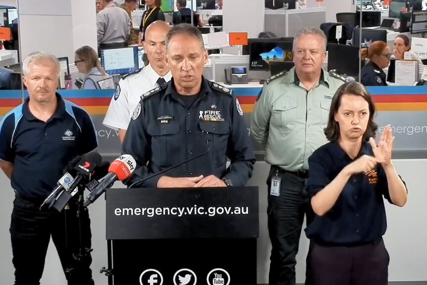 A man in the black uniform of a senior CFA manager speaks at a podium with a translator and three other emergency managers.