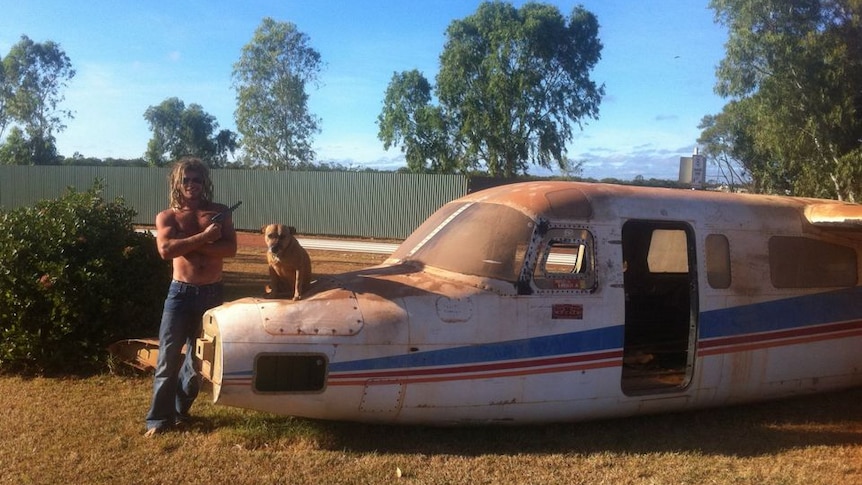 A man stands shirtless with a gun in one hand next to a dog sitting on an old airplane body.