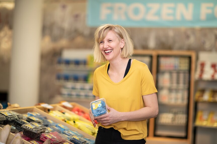 A smiling woman in a yellow tee-shirt in a grocery store aisle, holding a plastic punnet of faux blueberries.