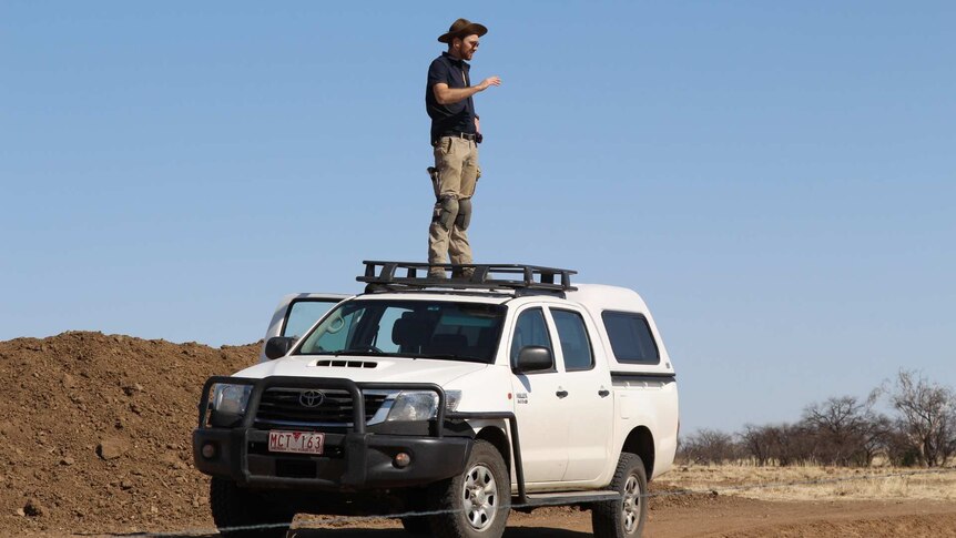 A man wearing dusty clothes, knee pads and a broad-brimmed hat stands on the roof of a four-wheel drive in the outback.