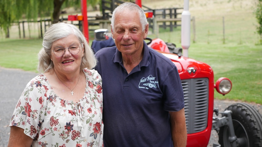 An elderly couple stand in front of a red tractor with grass and trees in the background 
