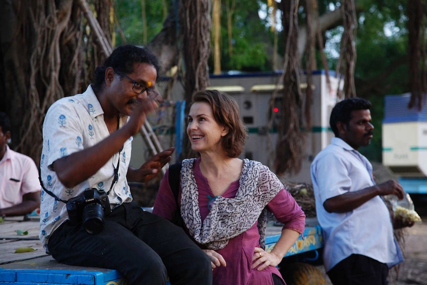 A man with a camera slung around his neck describing a shot while a woman in a sari listens intently