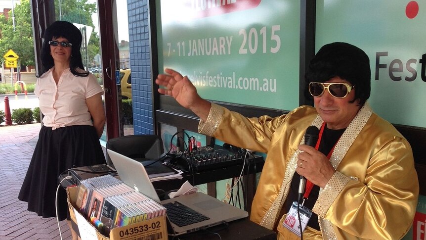 On a street corner an Elvis impersonator with a microphone sitting and a Priscilla Presley look-a-like next to him