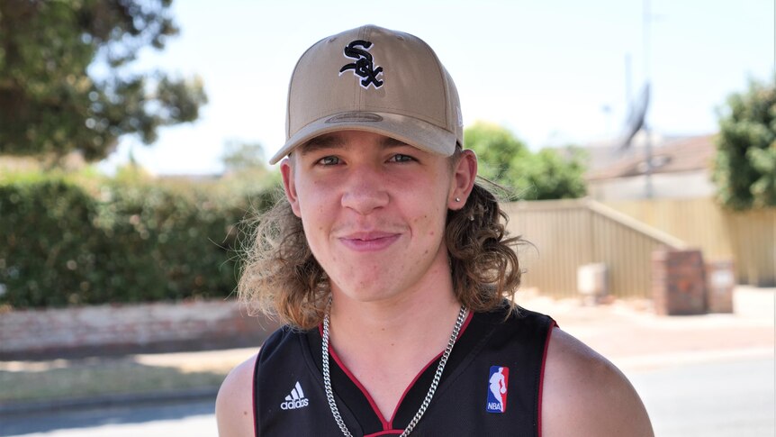 A teenager with a blonde mullet and a cap smiles at the camera with a suburban street pictured in the background.