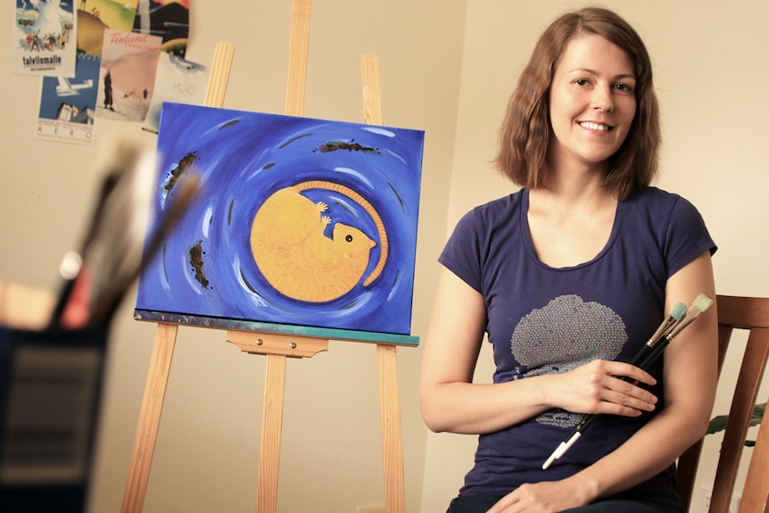 Ella Pesonen sits next to an easel holding paint brushes. The easel holds an artwork of an extinct rodent.