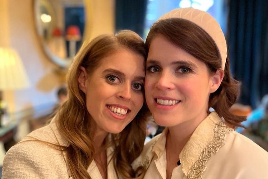 A close up of Princess Beatrice and Eugenie dressed in white jackets hugging each other and smiling.