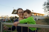 Woman holding two-year-old boy wearing a green jumper stands at a gate outside a house, smiling.