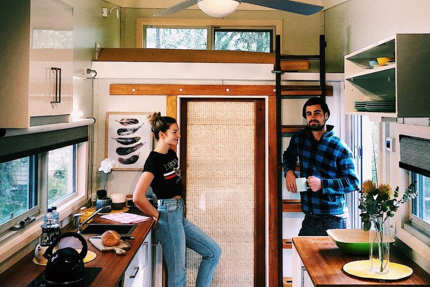 A young couple inside a tiny house that looks about the size of a large caravan. A ladder leads to a loft bed.