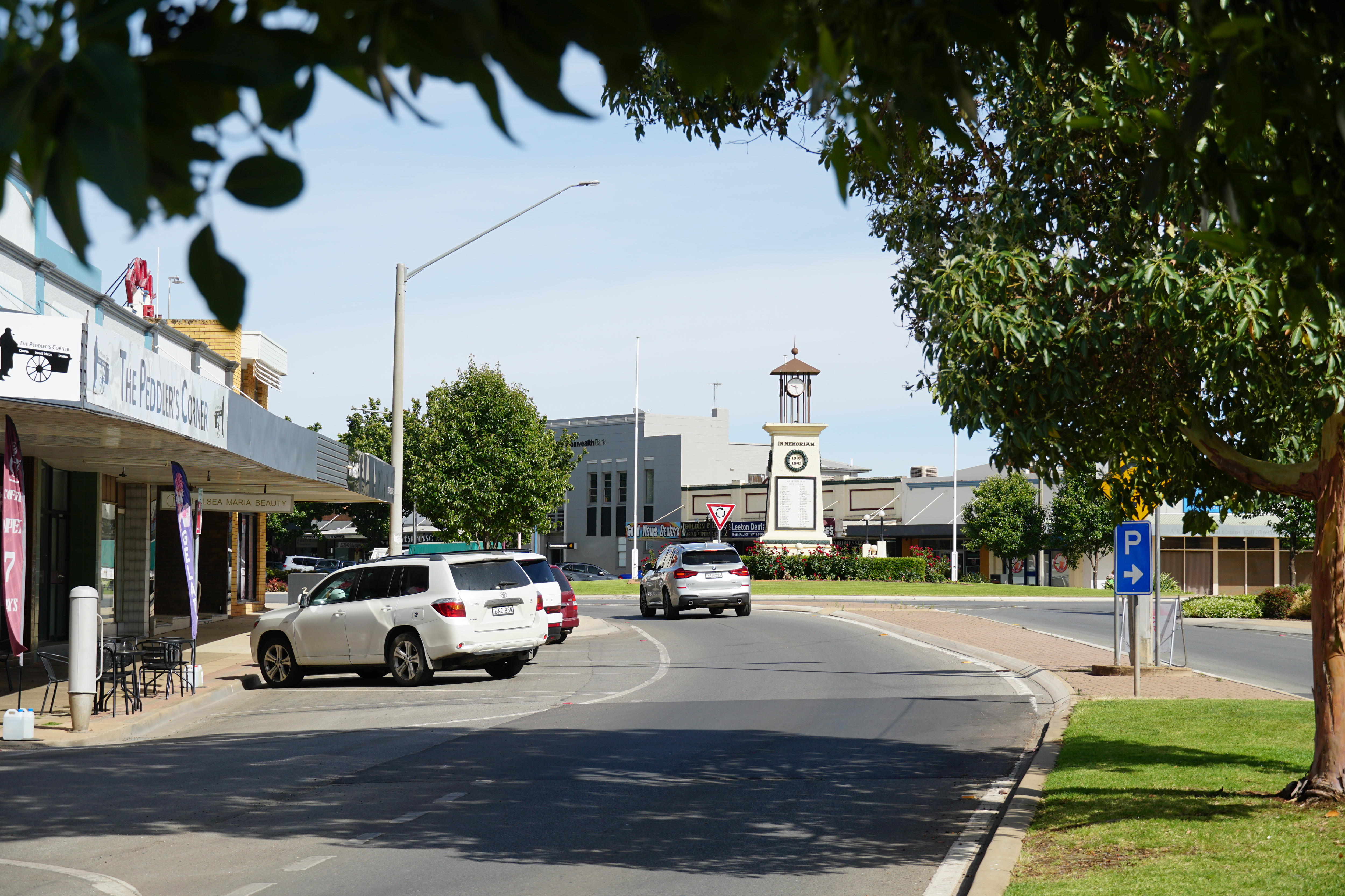 A street with a cafe, parked cars, and a car approaching a roundabout with a war memorial in its centre.