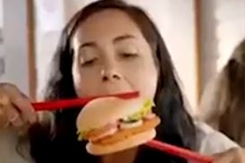 A dark-haired woman trying to eat a burger with giant red chopsticks