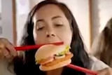 A dark-haired woman trying to eat a burger with giant red chopsticks
