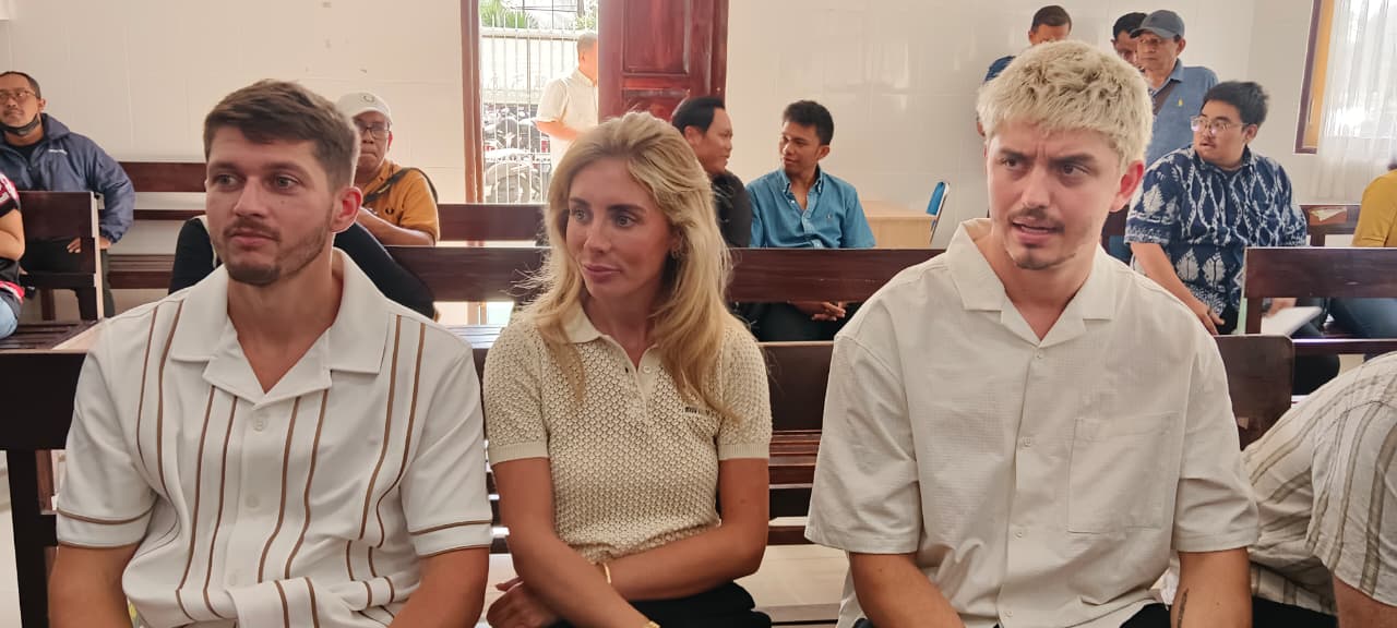 A blonde woman sits between two young men on a pew in a courtroom.
