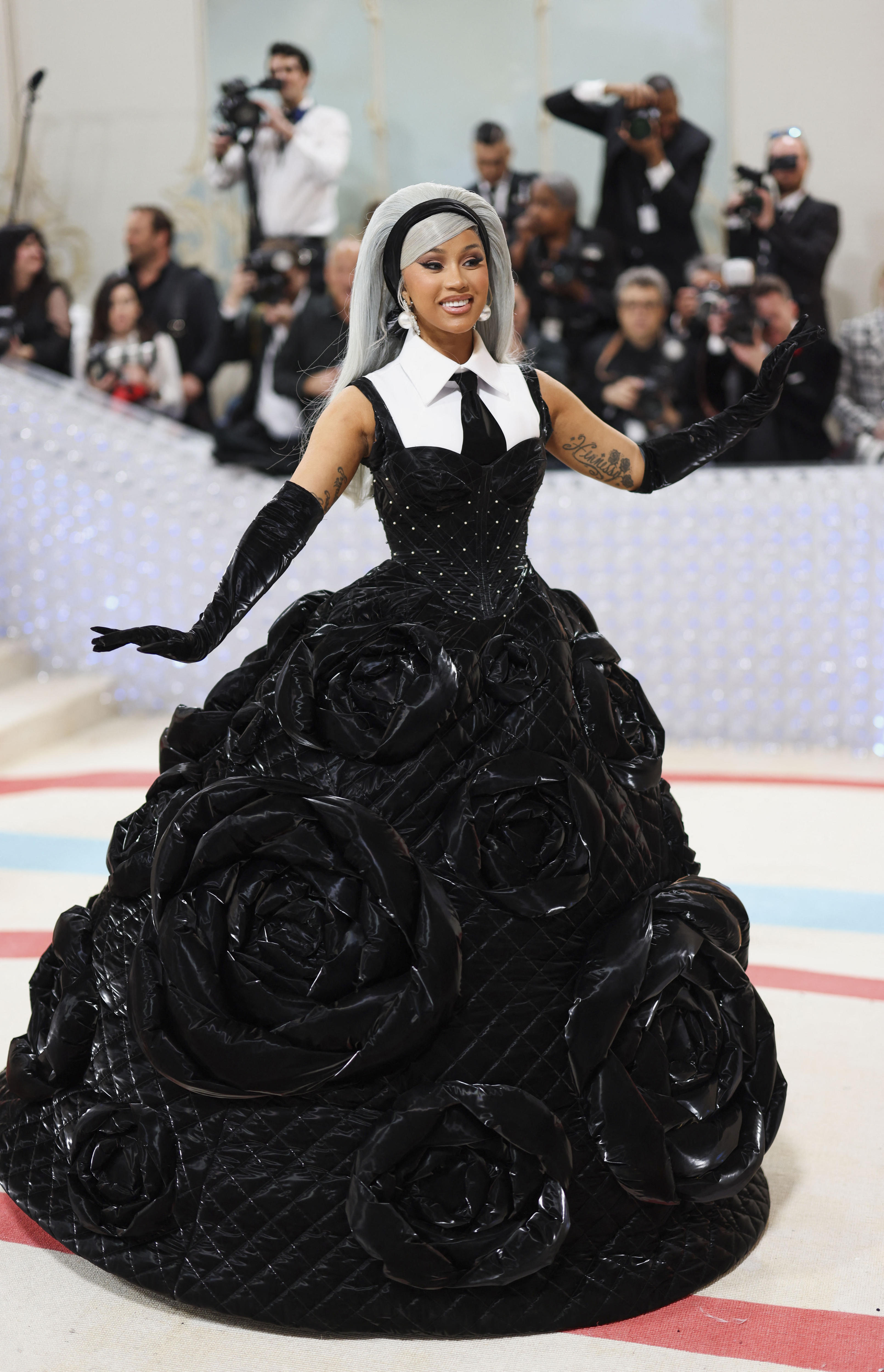 Cardi B in a black full-skirted cushion-gown with big plastic roses and a white sleeveless shirt with a black tie and grey wig