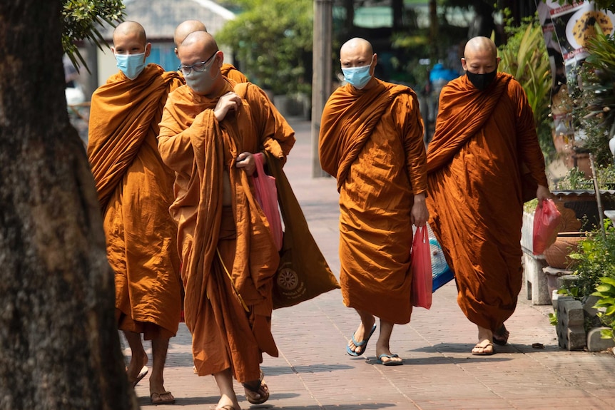 A group of men in orange robes and wearing masks walk along a tree-lined street.