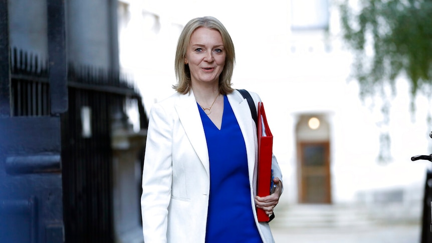 A woman in a blue dress and white coat walks down a grey street carrying a red folder.