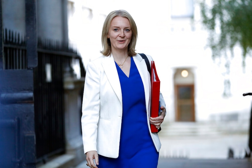 A woman in a blue dress and white coat walks down a grey street carrying a red folder.