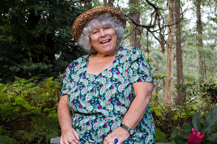 A photo of Miriam Margolyes who is wearing a bright patterned dress and sitting in a bush setting