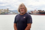 A well-built man with long, curly hair standing in front of a harbour.