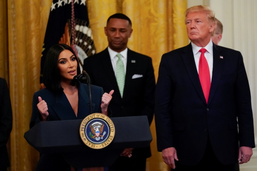 Kim Kardashian West speaks behind a lectern with a US presidential seal as President Donald Trump watches on.