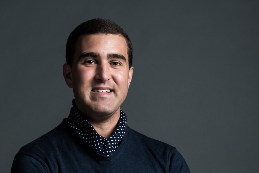 A man wearing a button up shirt smiles for the camera against a grey background