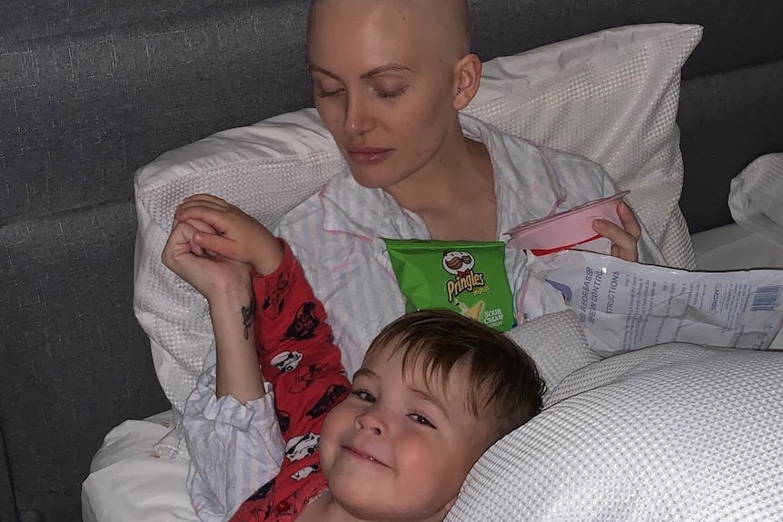 A bald woman laying in a bed with a young boy, holding hands.