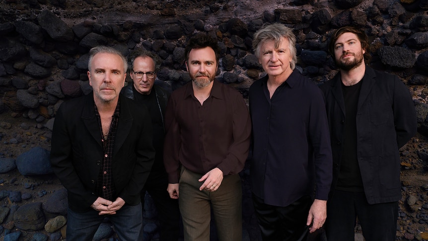 Five members of Crowded House stand in a line looking at the camera. A rocky hill is behind them.