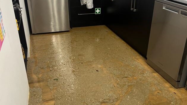 a kichen with brown cupboards and silver fridge with wooden floor covered in brown liquid 