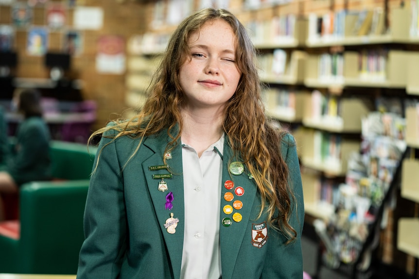 Abi wearing a green formal school jacket adorned with different badges, and smiling in school library.