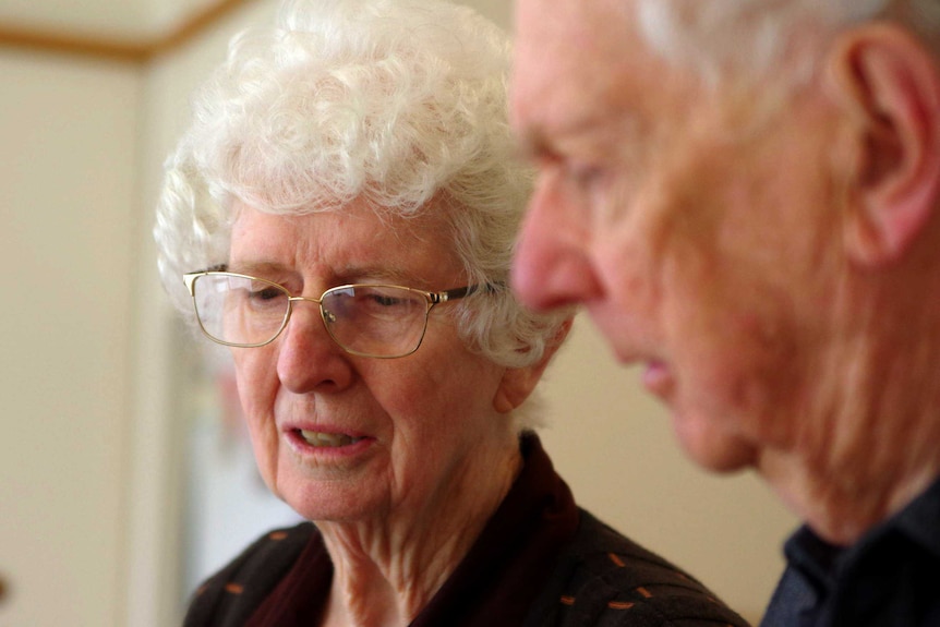 An elderly woman wearing glasses and her husband sit at a kitchen table.