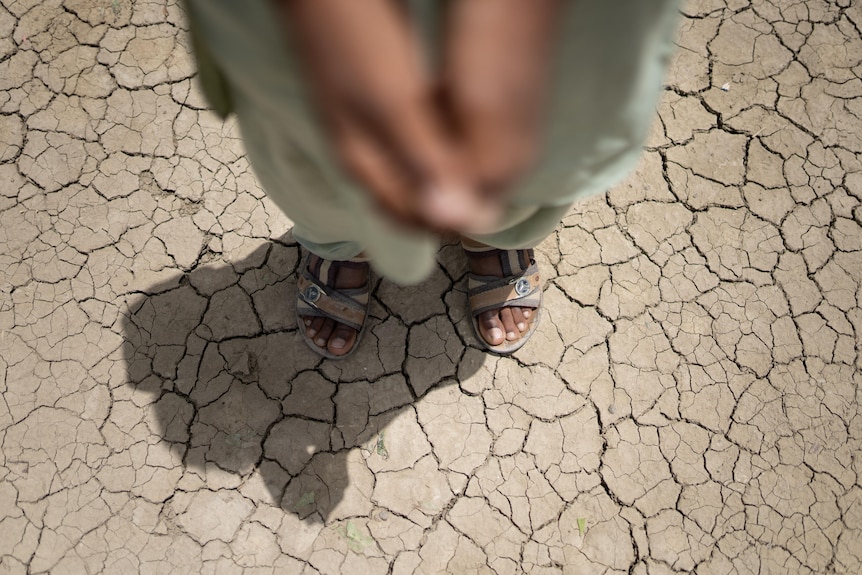 Person wearing sandals stands on dry, cracked earth. Only their leg, feet, and a blurred view of their hands are visible.