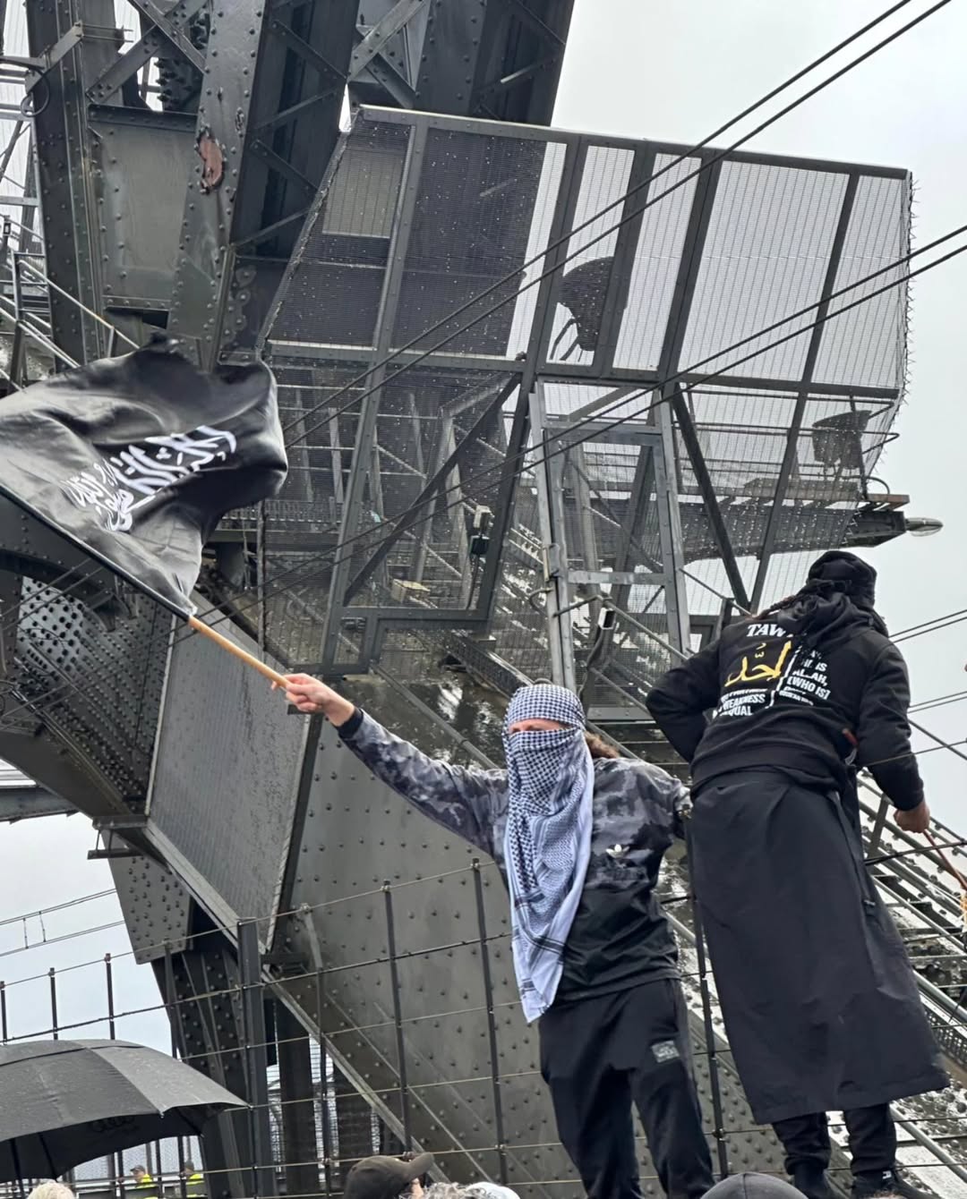 Um homem com o rosto envolto em uma roupa agita uma bandeira preta e branca da Shahada na Sydney Harbour Bridge.