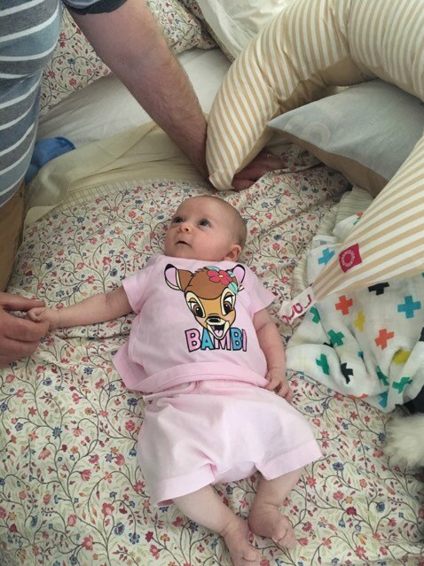 A newborn baby lies on a bed, looking up at the man sitting next to her.